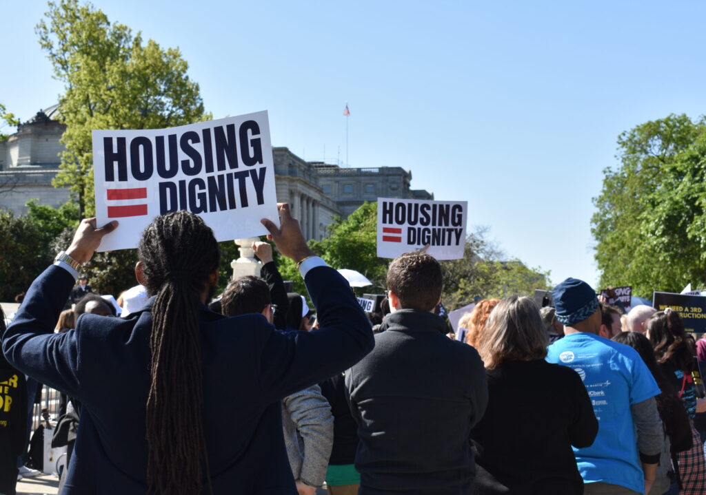 Housing = Dignity is on signs held by people gathered.